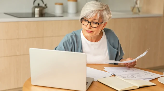 Une Femme Proche De La Retraite Calcule Sa Pension De Base Avec Une Calculatrice à La Table De Sa Cuisine.